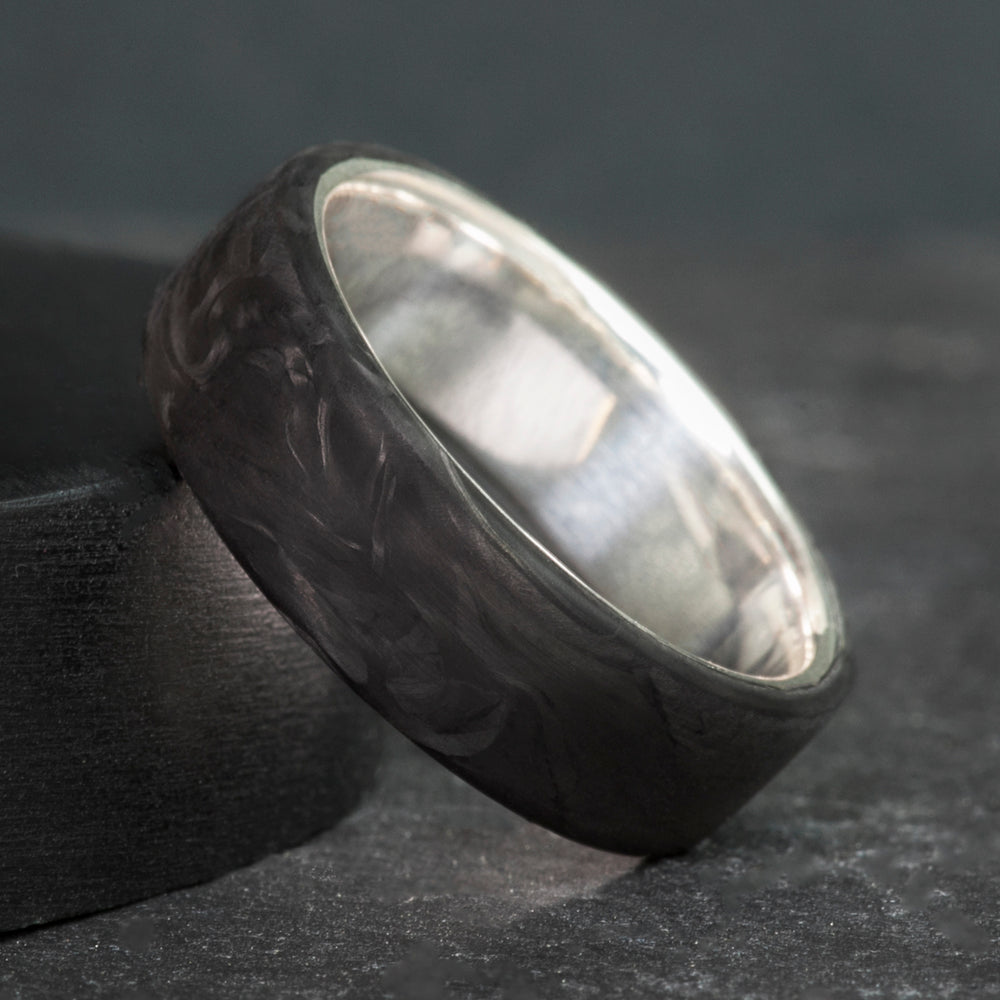 Black forged carbon fiber ring on a sterling silver sleeve, standing on dark slate