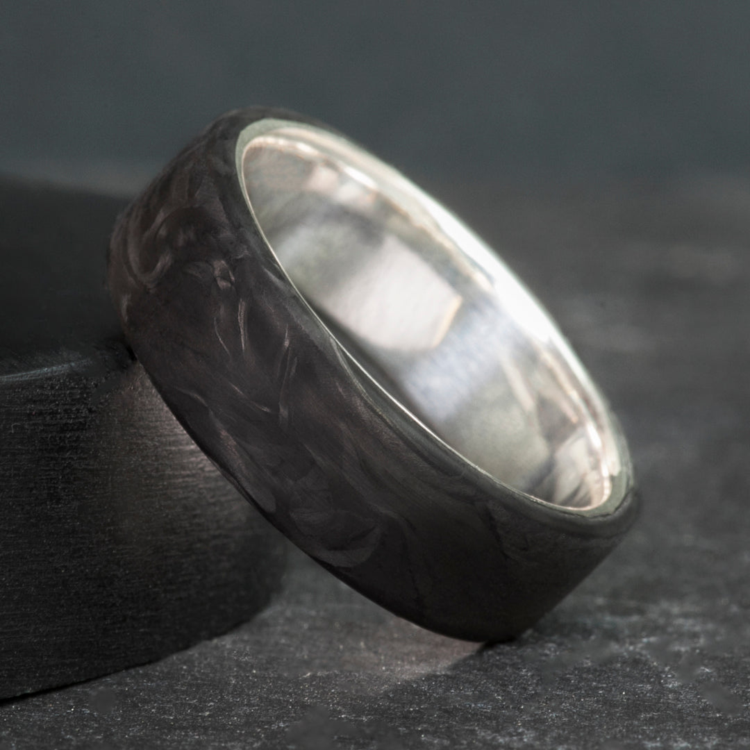 Black forged carbon fiber ring on a sterling silver sleeve, standing on dark slate