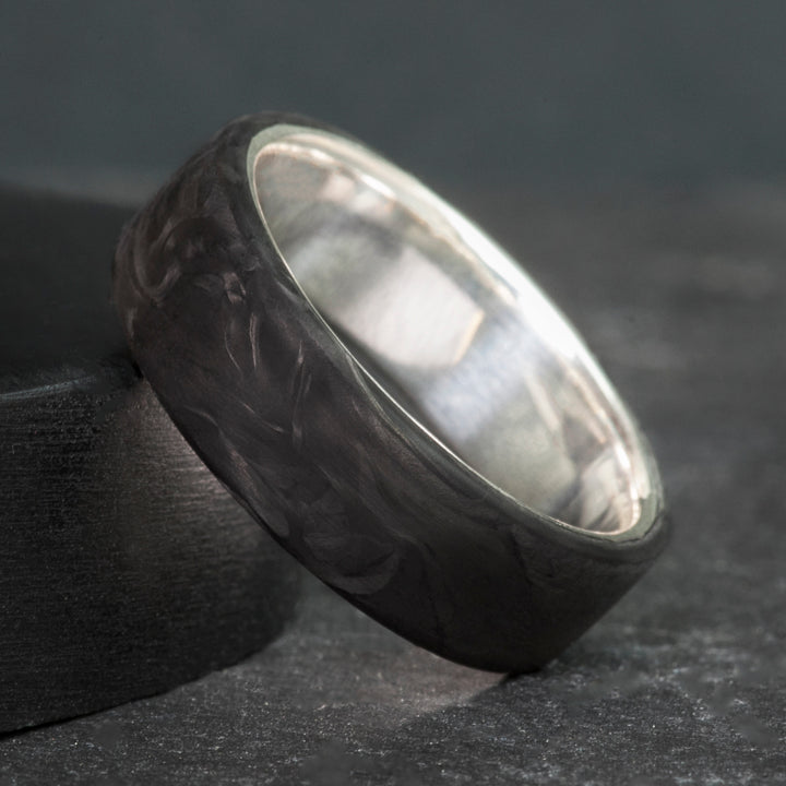 Black forged carbon fiber ring on a sterling silver sleeve, standing on dark slate