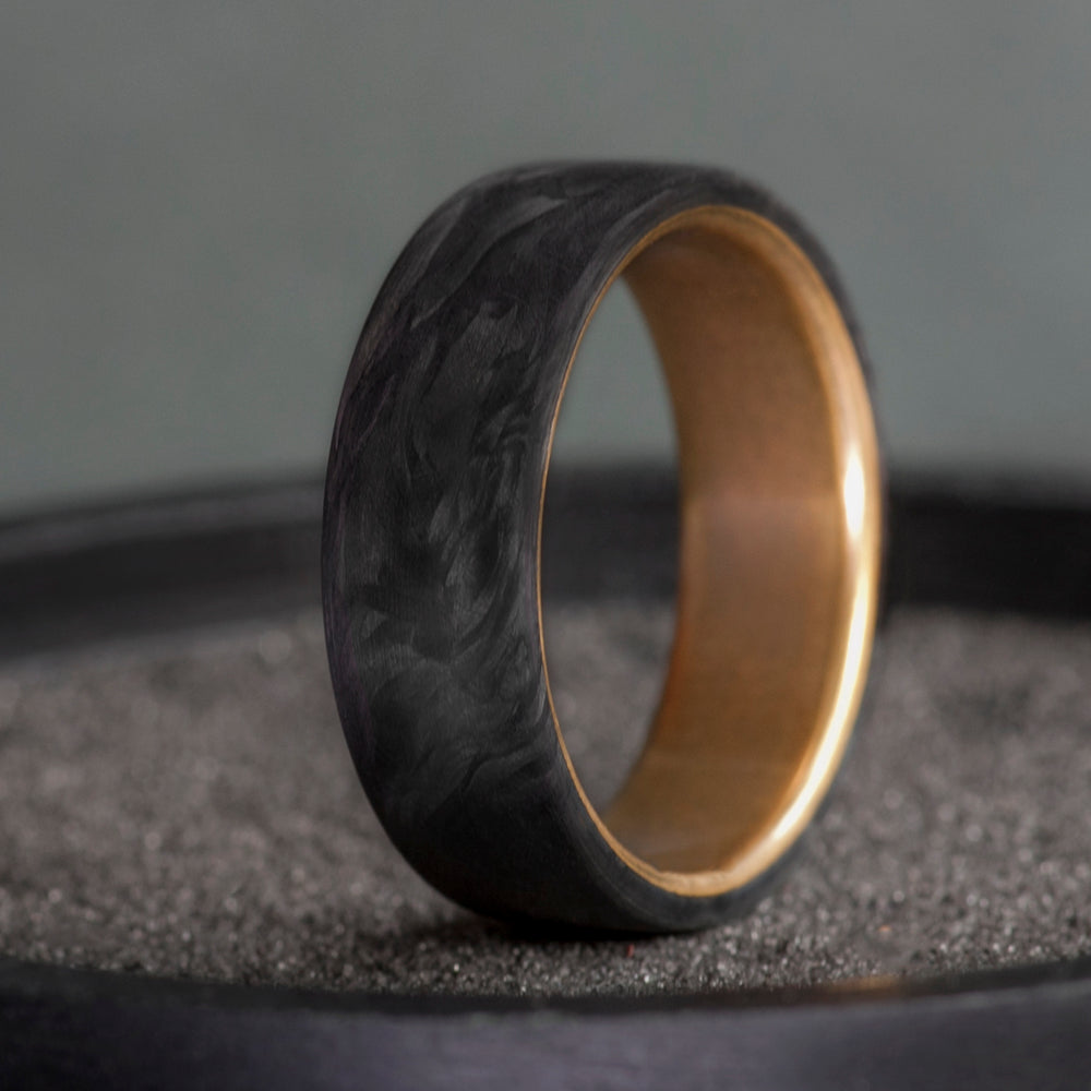 Black forged carbon fiber ring with warm brass interior on a dark textured surface