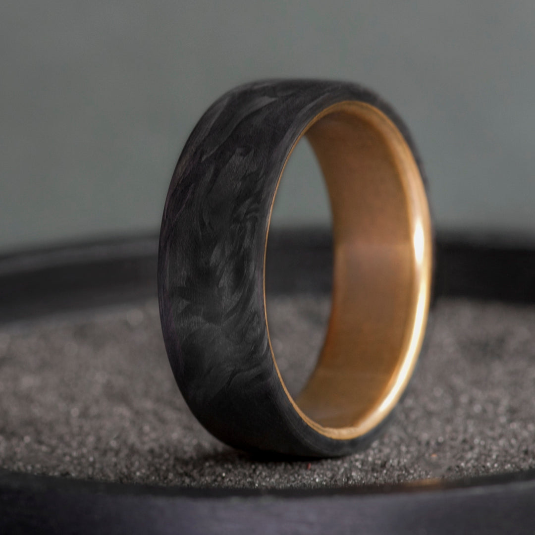 Black forged carbon fiber ring with warm brass interior on a dark textured surface
