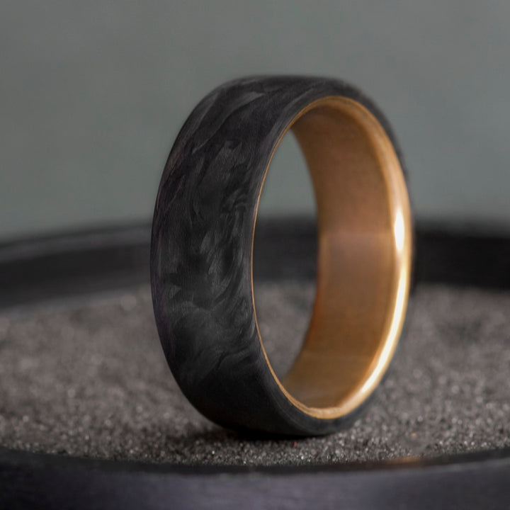 Black forged carbon fiber ring with warm brass interior on a dark textured surface